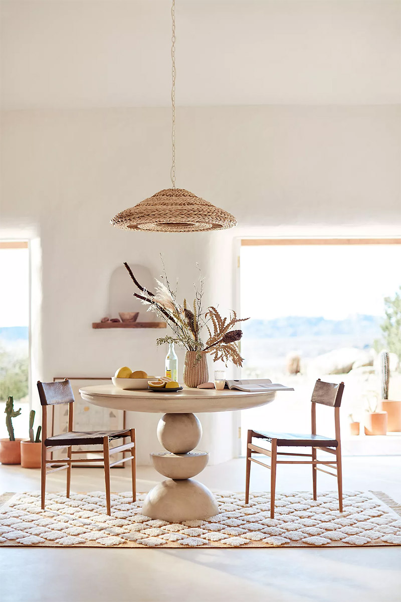 A rattan lamp hangs over a pedestal dining table flanked by two simple leather chairs.