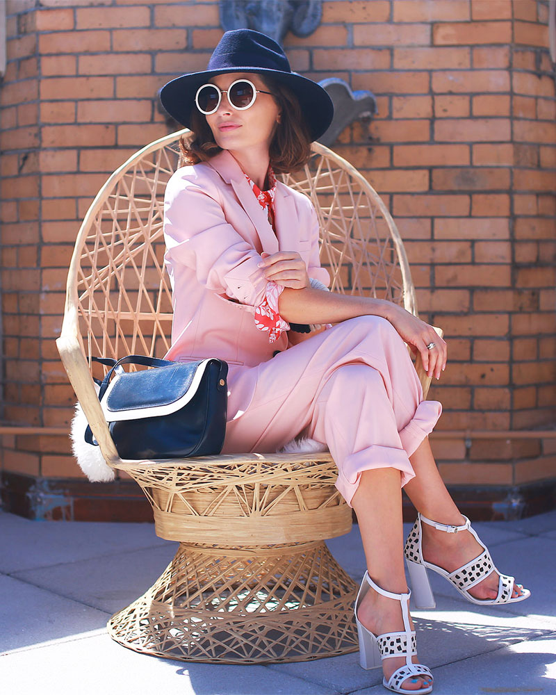 A woman in a dark blue hat poses in a peacock chair wearing a light pink pantsuit in New York City