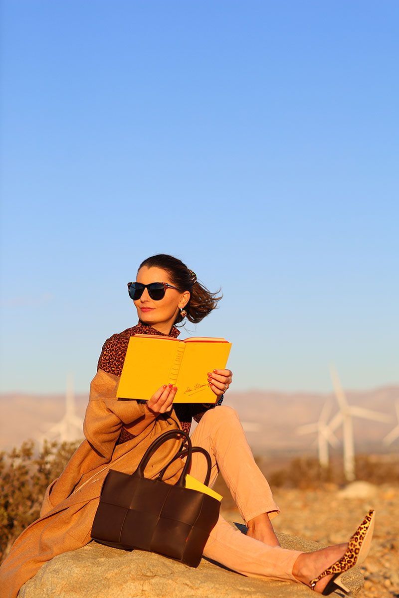 Fashion blogger Kelly Golightly wear a chic camel coat, pink velvet jeans and a leopard turtleneck and heels while sitting on a large rock in Palm Springs, reading a yellow book.