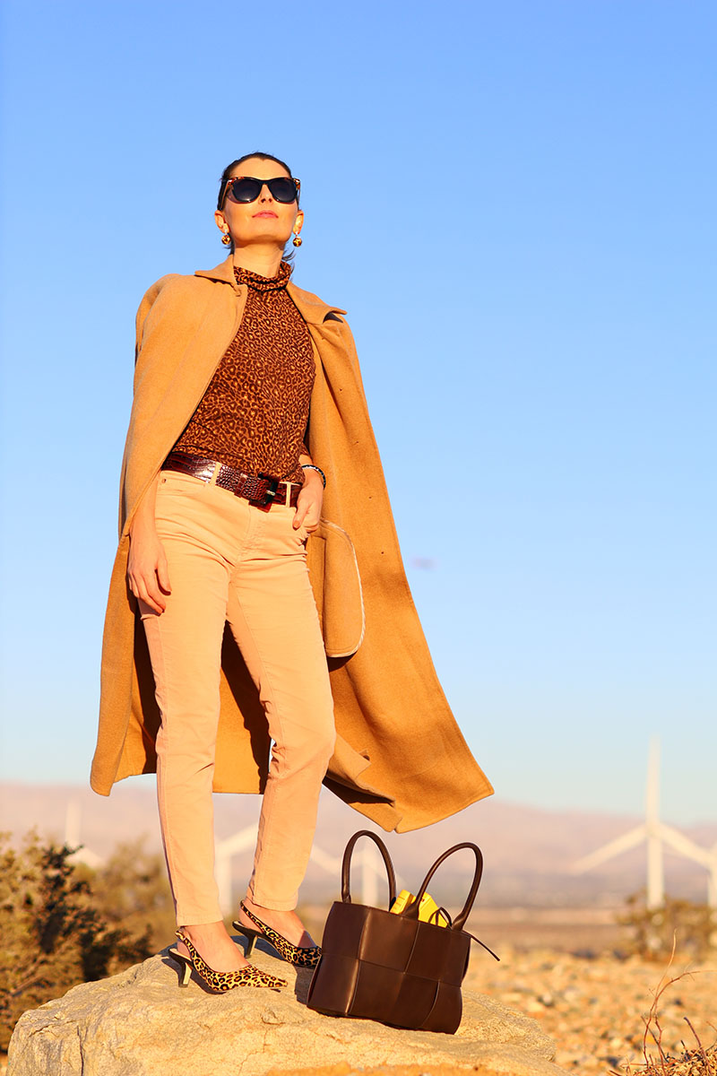 Fashion blogger Kelly Golightly wear a chic camel coat, pink velvet jeans and a leopard turtleneck and heels while standing in Palm Springs.