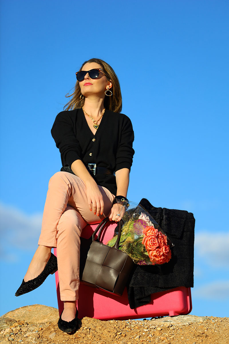 Fashion blogger Kelly Golightly wears pink velvet pants with a black cashmere sweater, while sitting on a vintage pink suitcase. A bouquet of roses rests in a dark brown leather handbag.