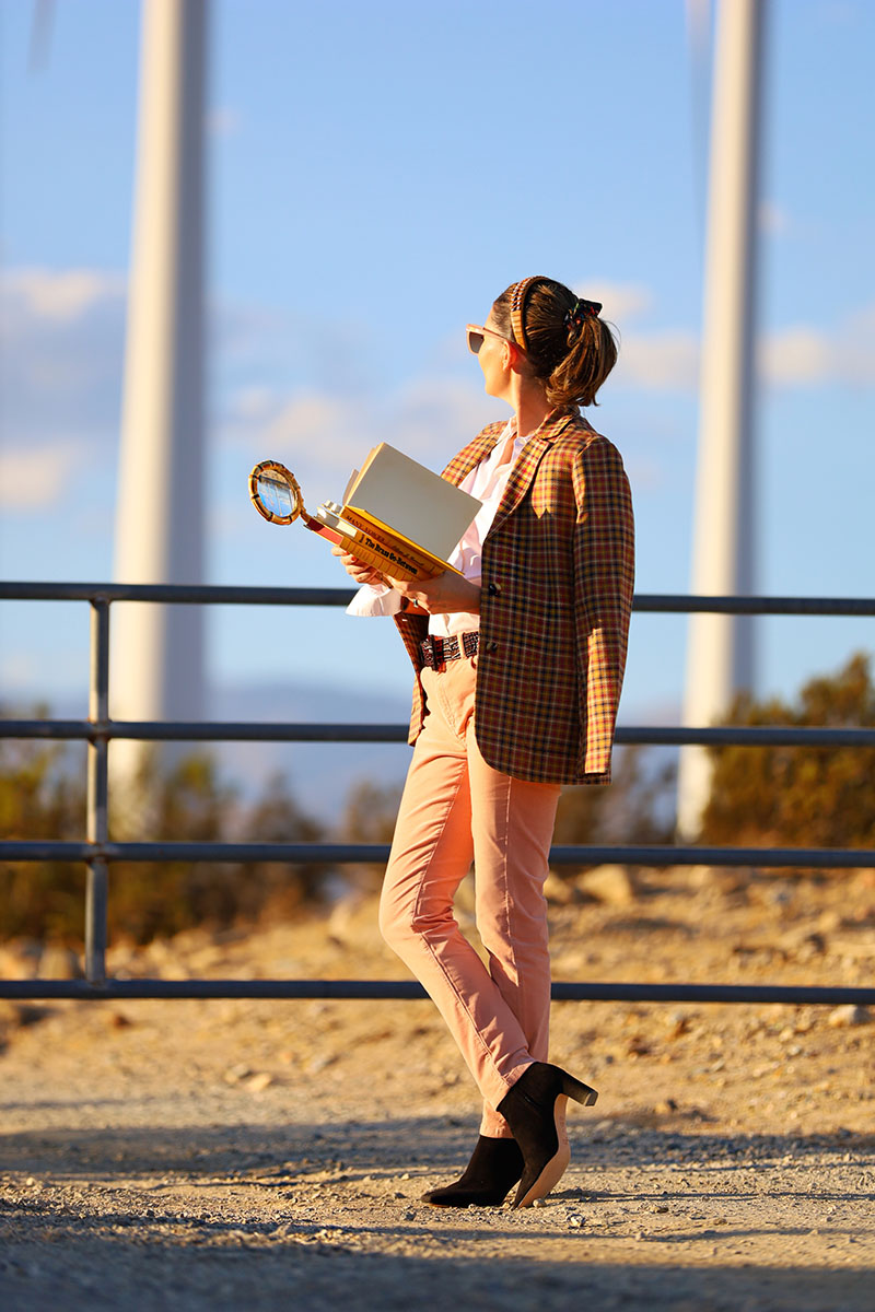 Fashion blogger Kelly Golightly wears a houndstooth blazer with pink velvet pants and a white button-down shirt. She holds a yellow book and a bamboo magnifying glass in her hand.