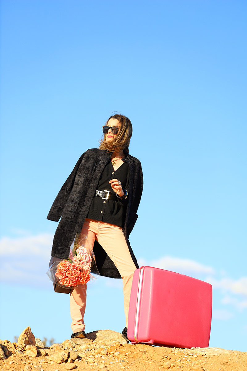 Fashion blogger Kelly Golightly wears pink velvet jeans pants with a black cashmere sweater and black faux fur coat draped over her shoulders. She completes the styling while holding a dark brown leather handbag filled with bouquets of roses.