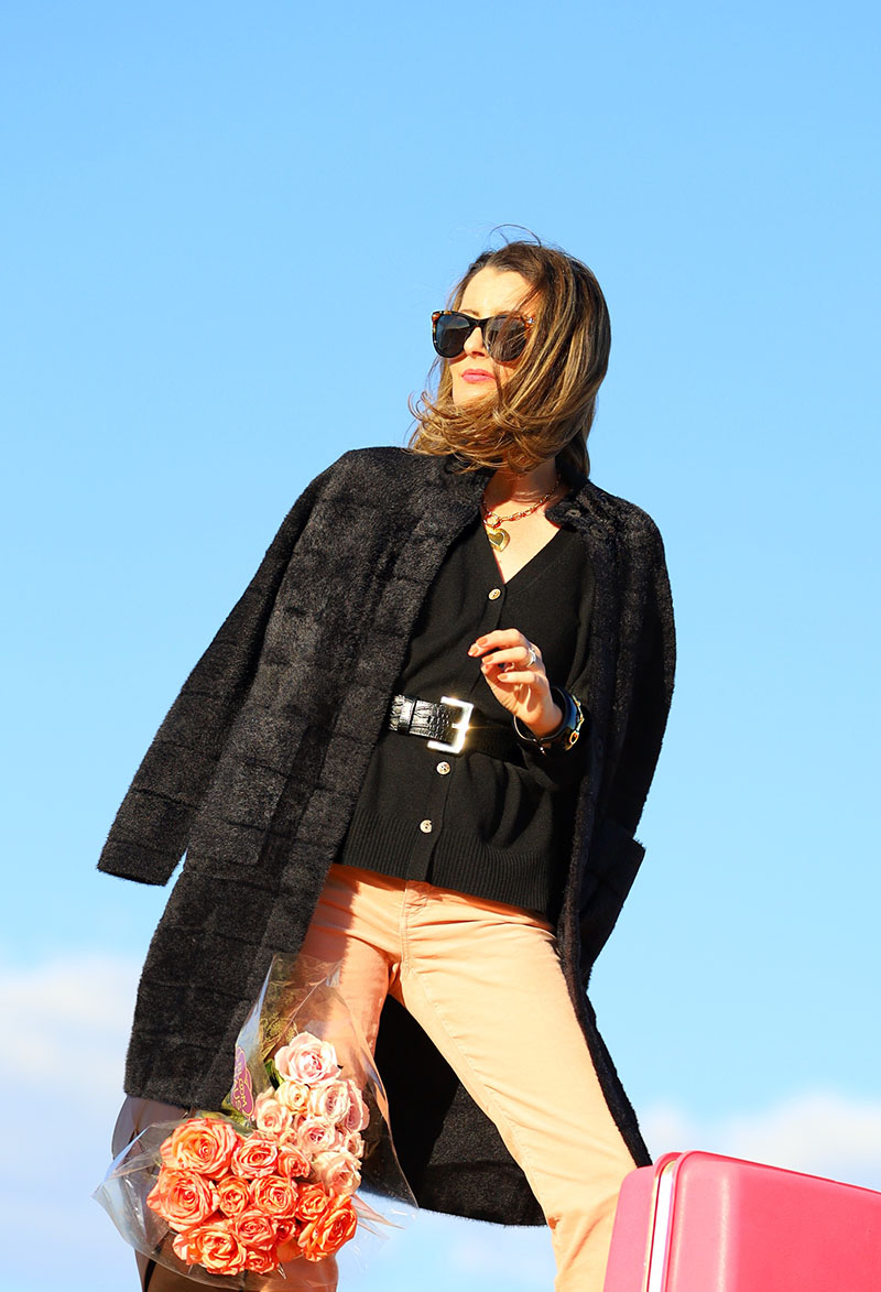 Fashion blogger Kelly Golightly is styling pink velvet pants with a black cashmere sweater and black faux fur coat while holding a dark brown leather handbag filled with bouquets of roses.