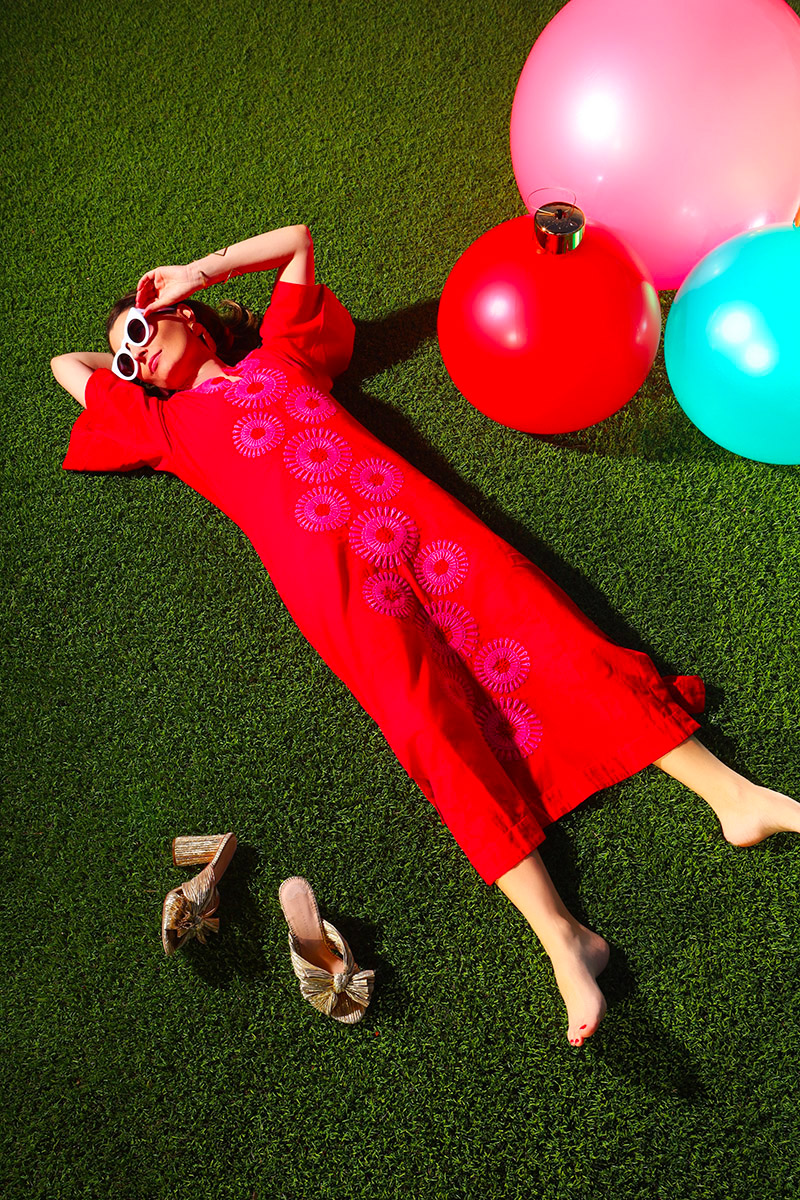Kelly Golightly wears a red and pink caftan while laying on artificial grass, next to giant inflatable ornaments.